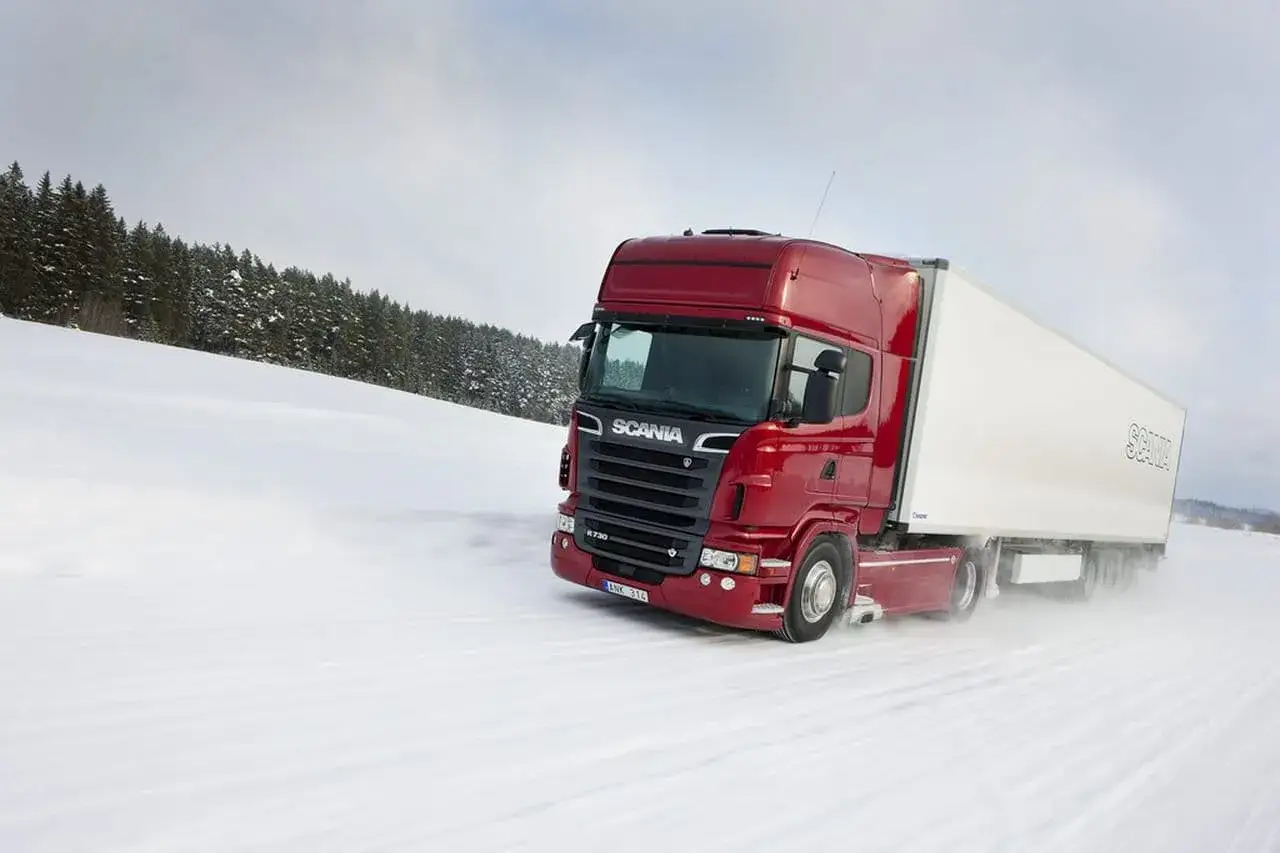 Camión Scania rojo con motor V8 avanza por un camino nevado, rodeado de árboles cubiertos de nieve.