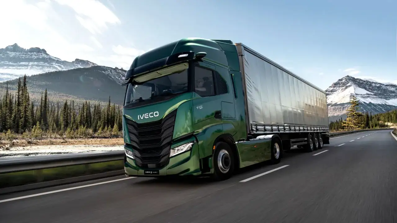Camión Iveco S-Way verde brillante recorre una carretera escénica rodeada de montañas nevadas y bosques.