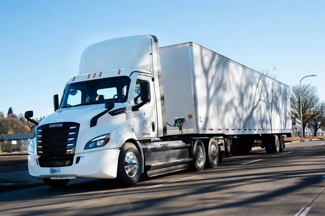 Camión eléctrico blanco de Freightliner, parte de la flota de Volvo Group, circulando por una carretera con el sol brillando.