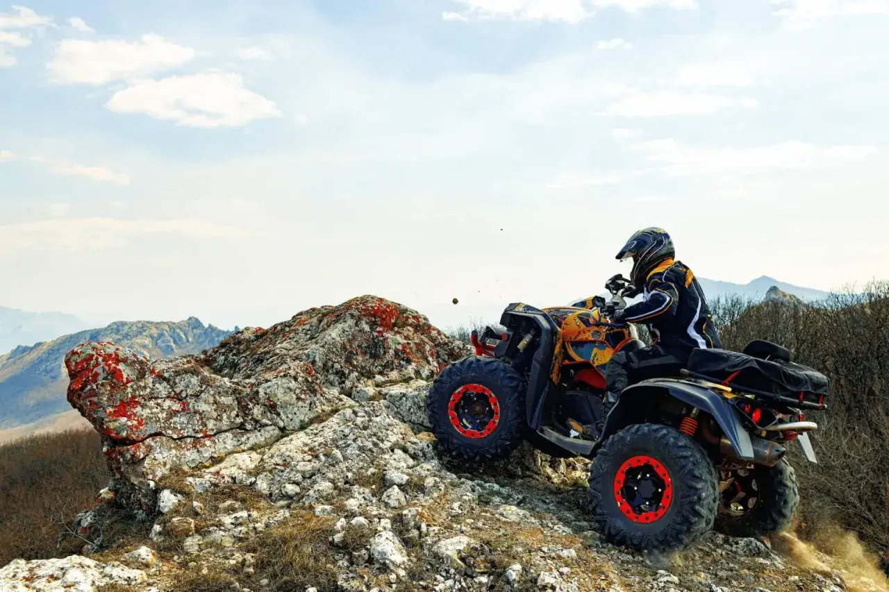 Un piloto con casco y chaqueta amarilla y negra conduce un quad ATV por un terreno rocoso y montañoso. El permiso para este tipo de aventura es esencial.