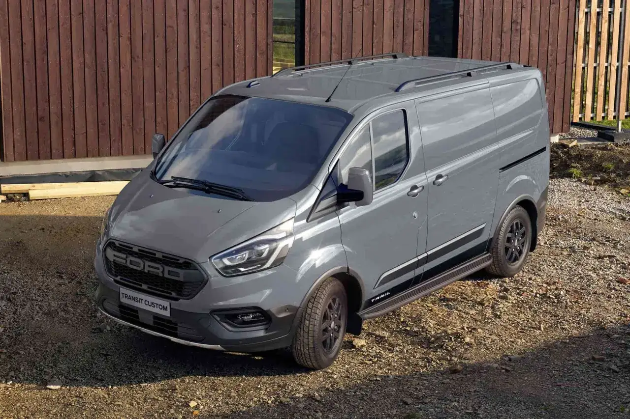 Furgoneta phev gris Ford Transit Custom Trail aparcada en grava, con vallas de madera de fondo.
