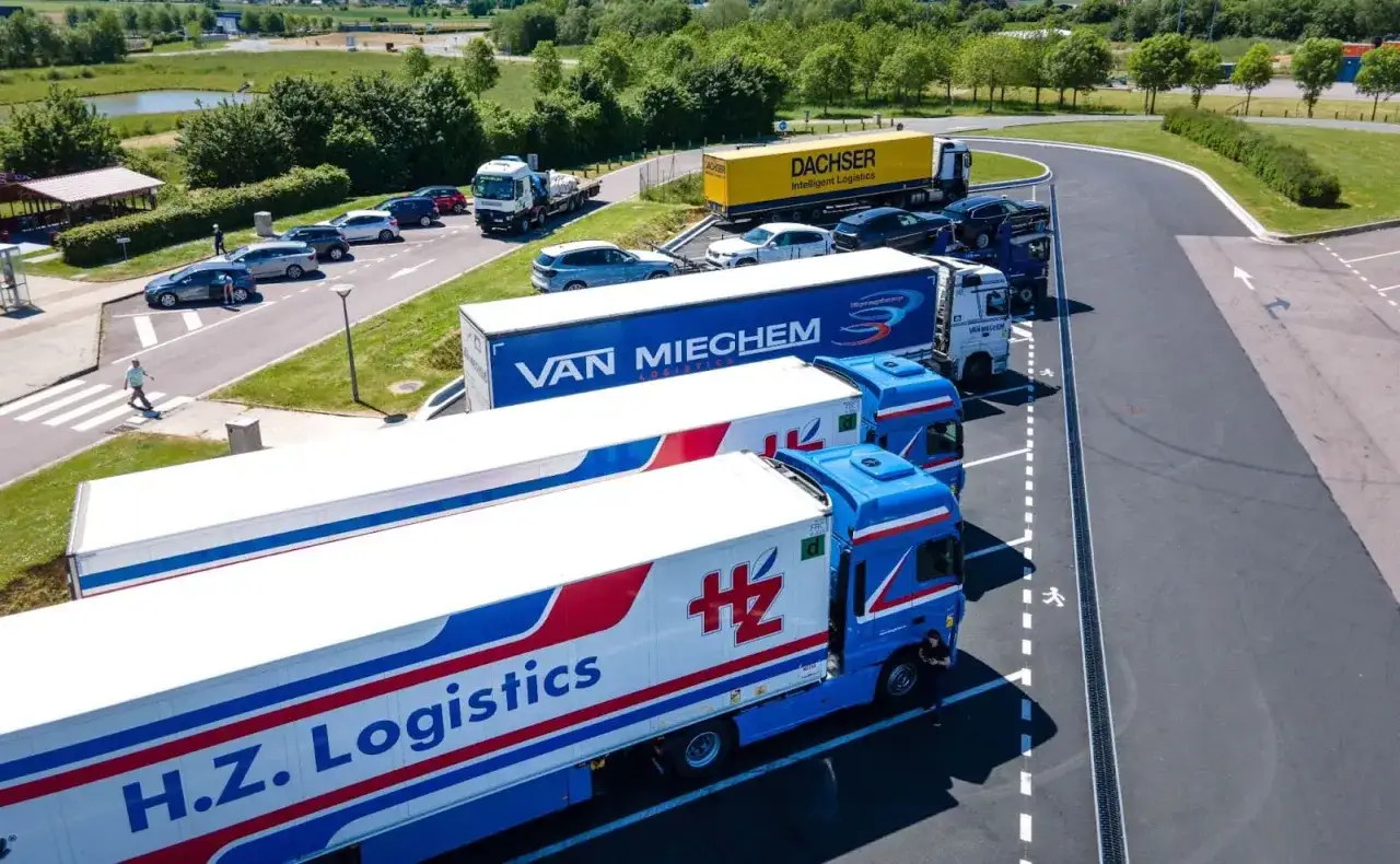 Varios tipos de camiones, incluyendo H.Z. Logistics y Van Mieghhem, estacionados en un área de descanso.