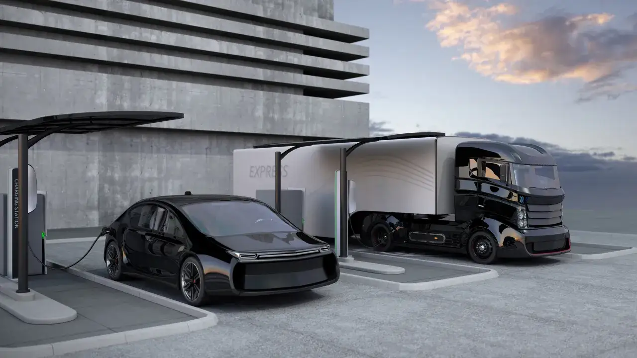Un coche negro y un camión del futuro cargando en una estación eléctrica frente a un edificio de hormigón.